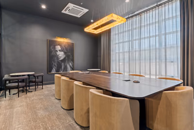 Dark grey board room with a long walnut table, tan upholstered chairs, a framed black-and-white portrait, a glowing rectangular pendant, and sheer curtains over a tall window.