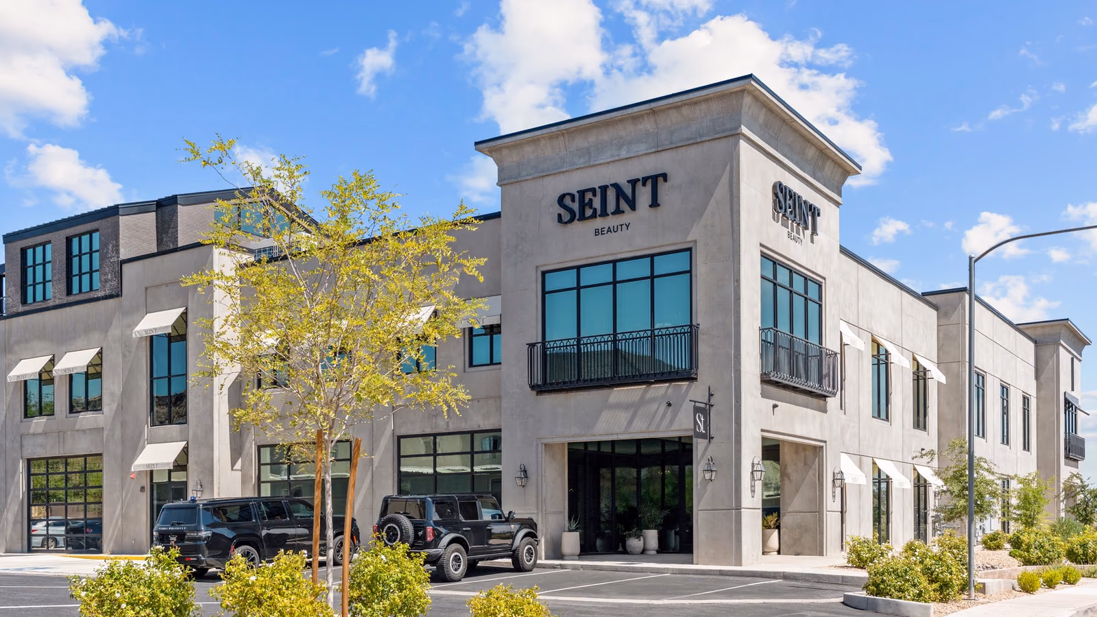 Two-story grey concrete commercial building with a tall corner tower carrying the Seint Beauty sign, black-framed windows, white awnings and a young tree in front, under a blue sky.